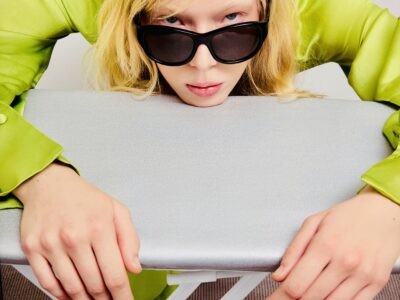 Model in a lime green satin shirt wearing black Blue Elephant sunglasses, leaning over an ironing board